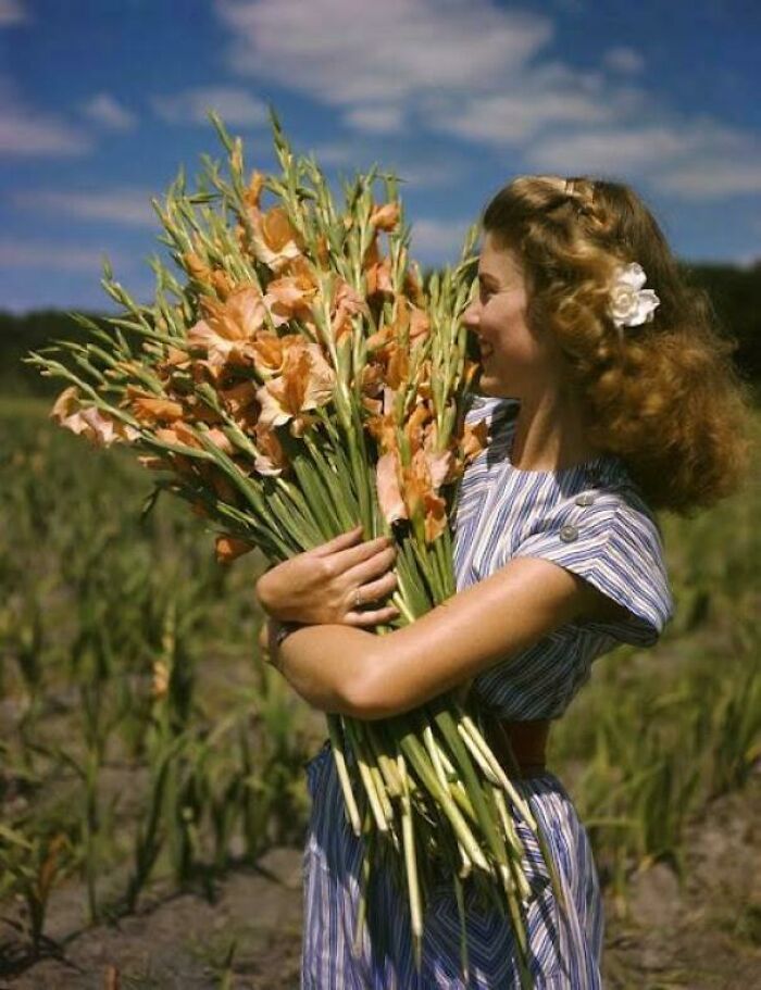 Young woman in a striped dress holding a large bouquet of flowers in a historic photo capturing life in a different era.