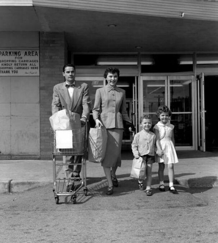 Family in vintage clothing walking with shopping bags and cart outside a store in a rare and fascinating historic photo.