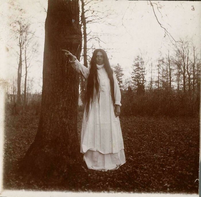 Woman in a long white dress with long hair standing in a forest, a rare and fascinating historic photo from a different era.
