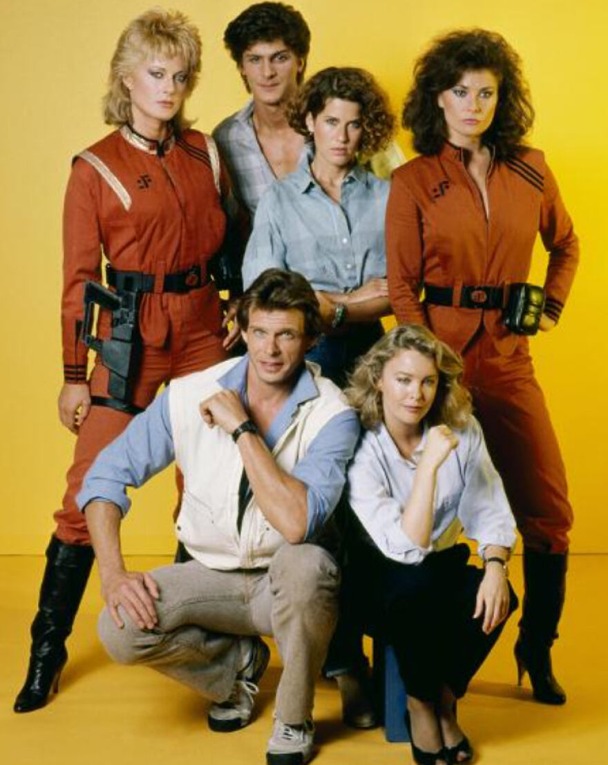 Group of six people in 1980s style clothing posing against a yellow background, showcasing iconic 80s fashion and culture.