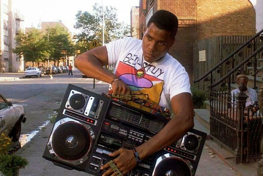 Man in 1980s style clothing carrying a large boombox on a city street, capturing a classic ‘80s lifestyle moment.