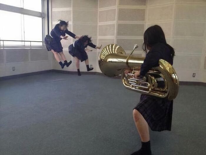 Three girls in school uniforms pulling off creative poses with a tuba in an empty, well-lit room.