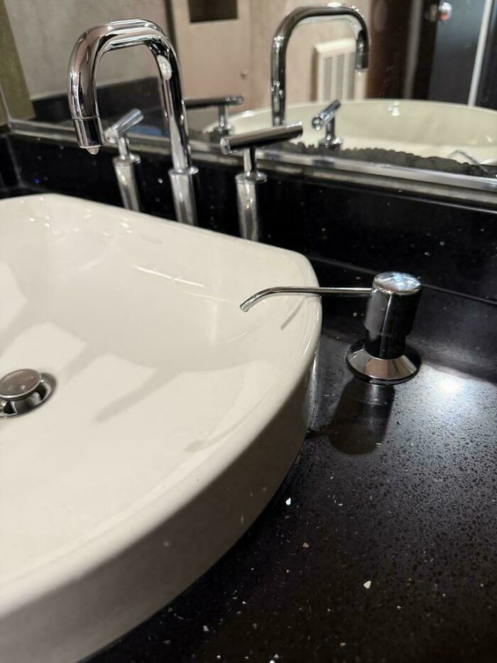 A bathroom sink with a faucet over the basin, and a soap dispenser comically positioned outside the basin, a funny You Had One Job fail.
