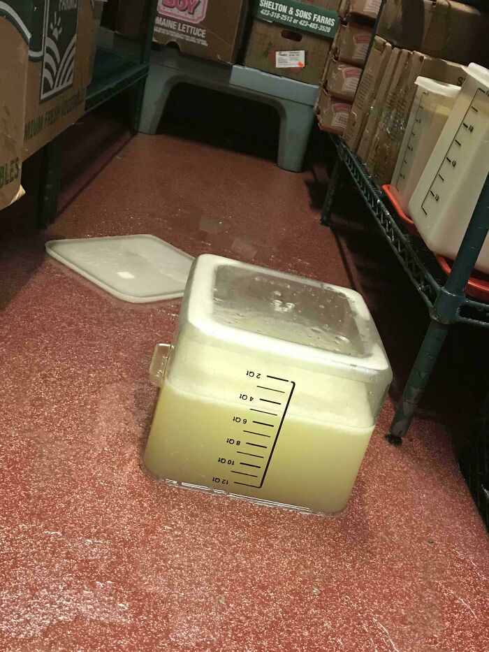 A container of liquid is lying upside-down on a wet, red floor with a lid nearby. This messy scene needs to be fixed.