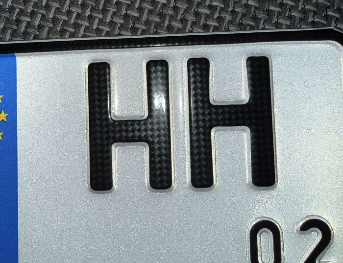 Close-up of a silver license plate with black carbon fiber letters HH, displaying upside-down elements that might need fixing.