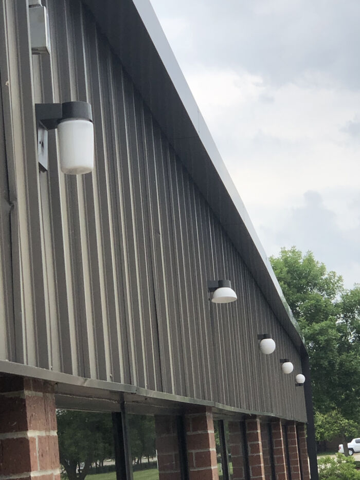 An upside-down light fixture on a gray corrugated metal wall, next to other outdoor lights, with brick below.