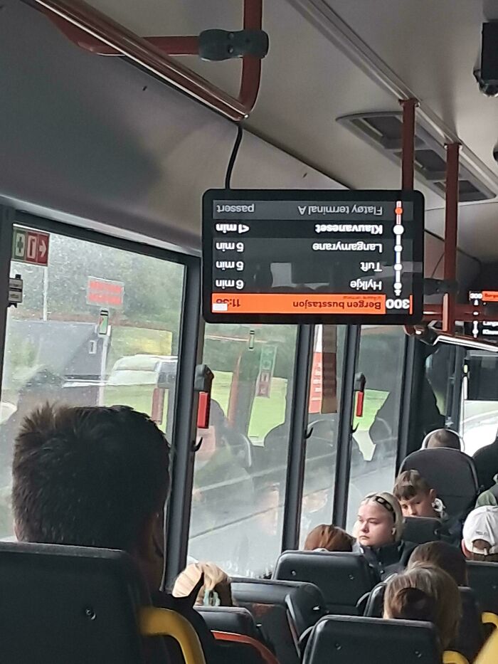An upside-down bus display showing upcoming stops and times, with passengers looking out the window, on a rainy day.