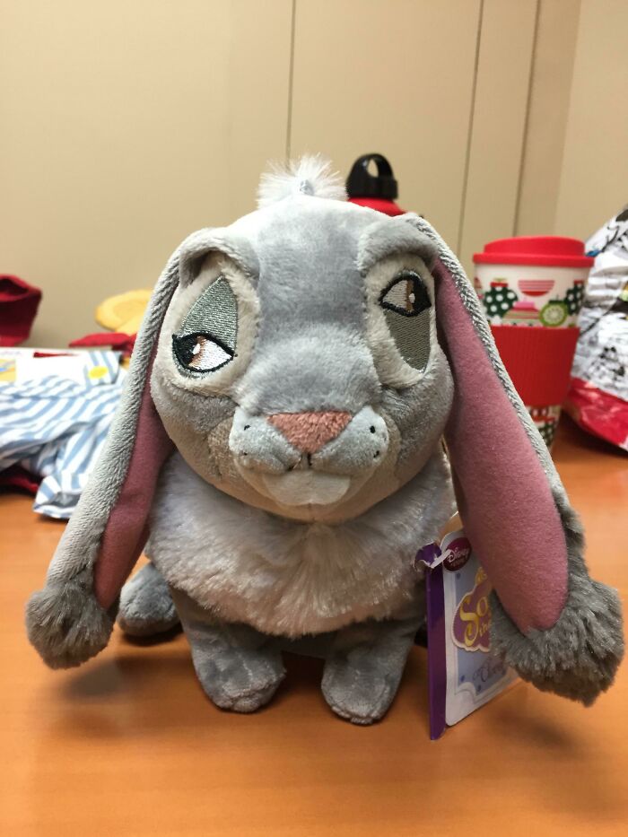 A gray rabbit plushie with long floppy ears sits on a brown desk, next to a red coffee cup. This upside-down plushie might need fixing!