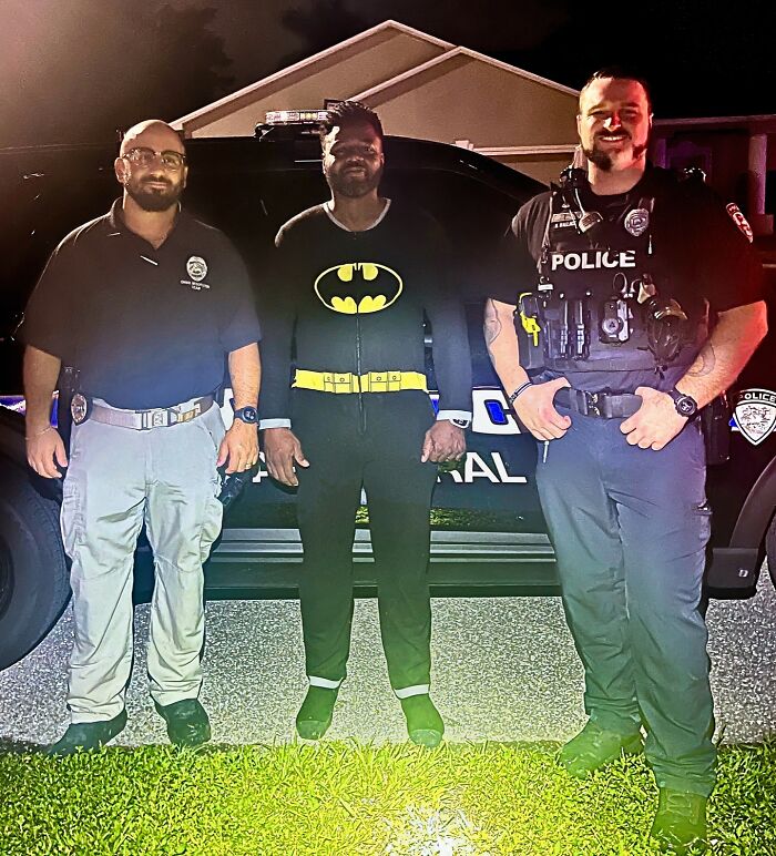Three men, including two police officers and one in a Batman outfit, standing by a police vehicle at night, showing gut warnings.