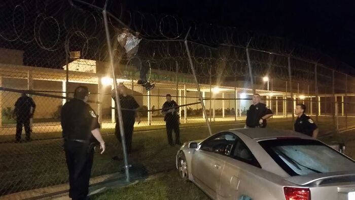 Person climbing barbed-wire fence at night while police officers watch, capturing gut warning and horrible event.