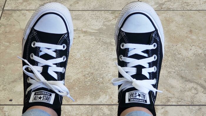 A person wearing black Converse All Star sneakers with white laces, viewed from above. The shoes are deliberately Upside-Down.
