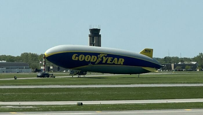 Goodyear blimp parked on an airport runway with control tower and buildings in the background, reflecting made up but real stories.