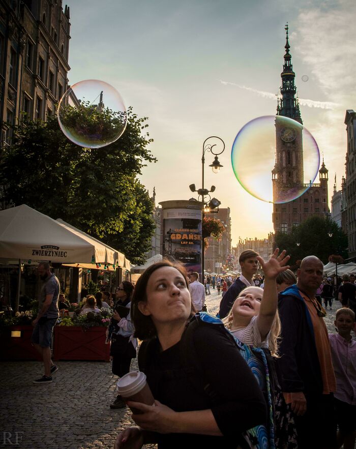 A young child reaches for a giant bubble in a town square, displaying an Accidental Renaissance aesthetic.