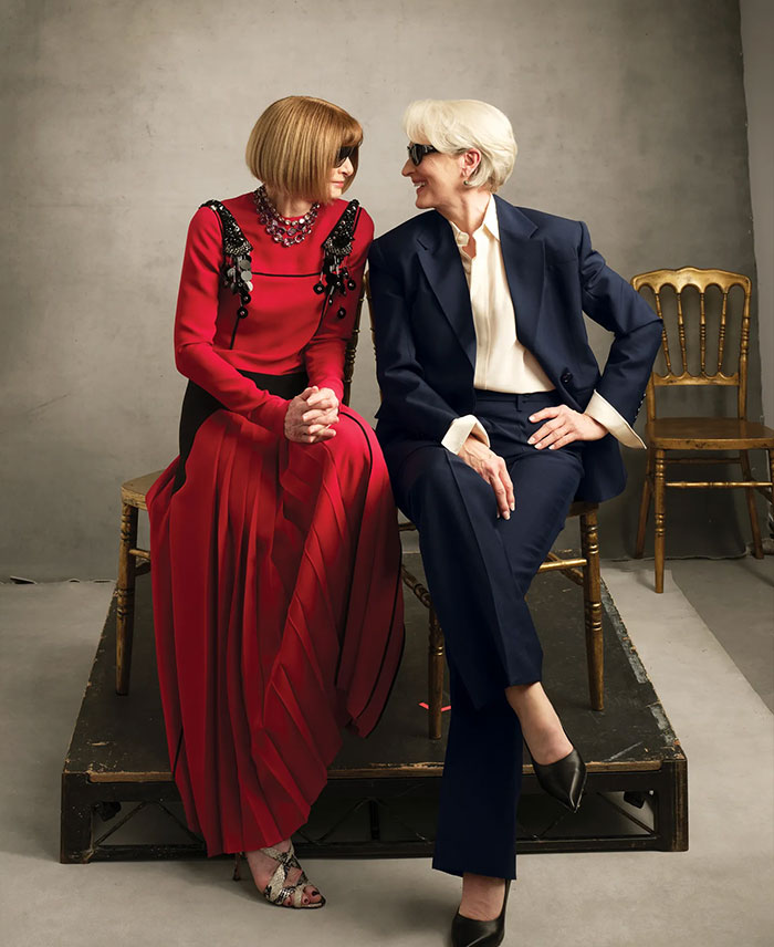 Two women wearing sunglasses and stylish outfits seated on chairs, representing Anna Wintour and Vogue fashion reality.