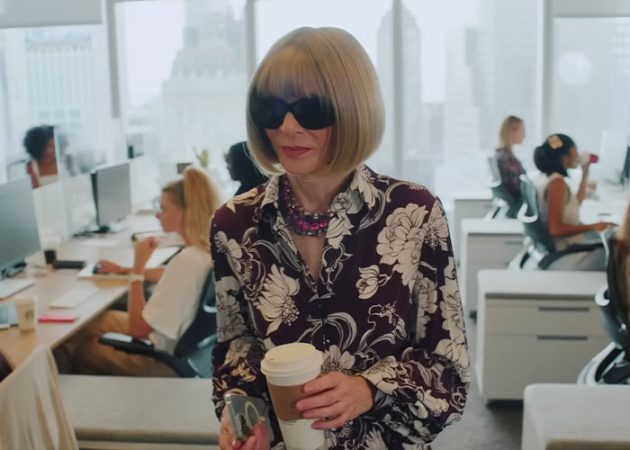 Anna Wintour wearing sunglasses and a floral blouse, holding coffee in a busy Vogue office with assistants at desks.