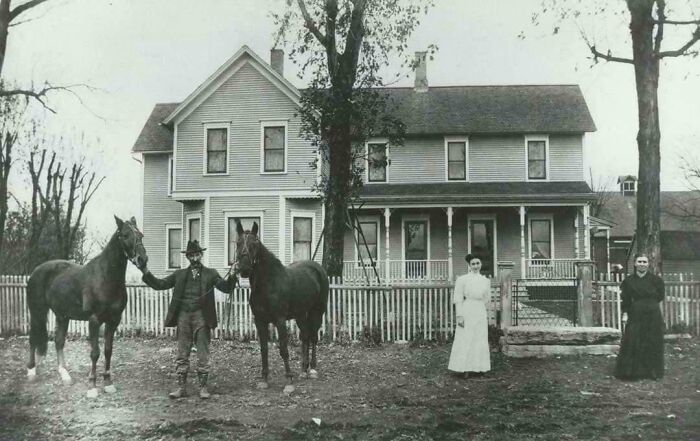 A candid old photo of a family, a man with two horses and two women, in front of a large house.