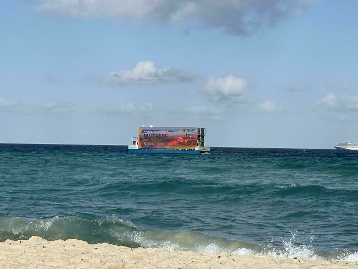 Floating digital billboard in the ocean off a sandy beach with waves under a partly cloudy sky showing dystopian United States scene