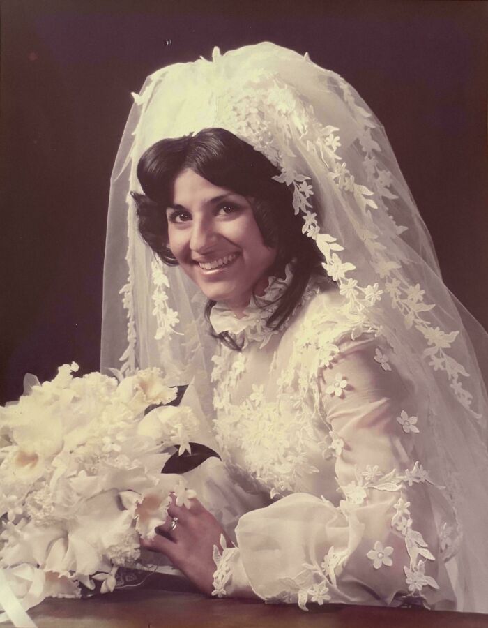 A candid old photo of a smiling bride in a white lace wedding dress and veil, holding a bouquet.