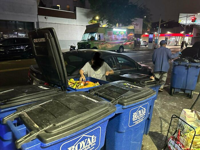Person scavenging food from a dumpster at night on an urban street, illustrating dystopian conditions in the United States.