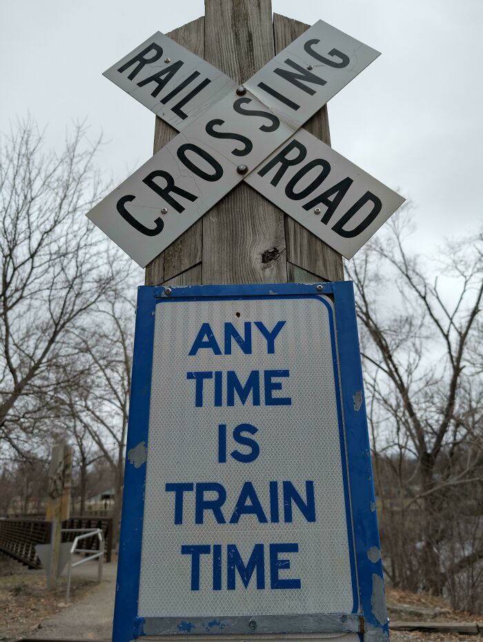 Railroad crossing sign with a humorous train time warning surrounded by bare trees on an overcast day.
