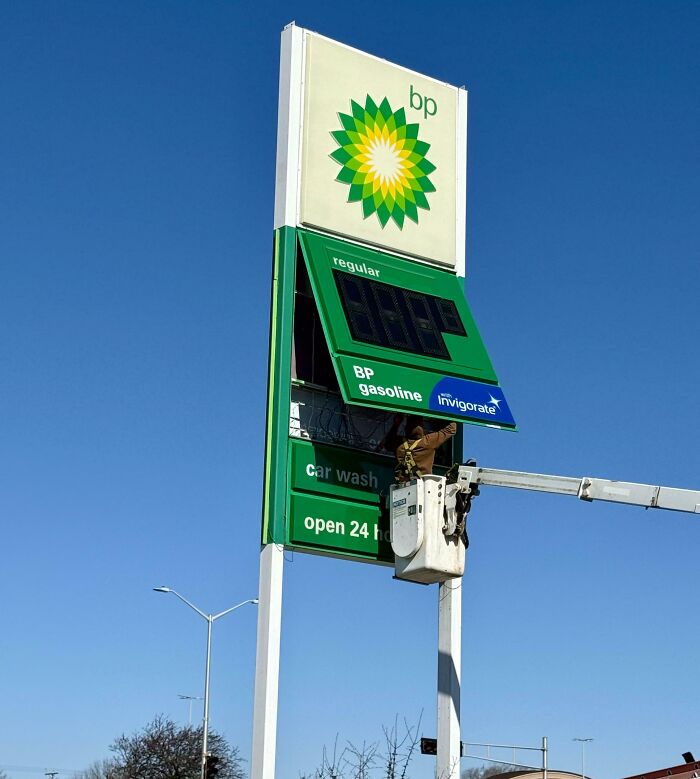 Worker in a bucket lift fixing a BP gas station sign under a clear blue sky, showing dystopian United States imagery.