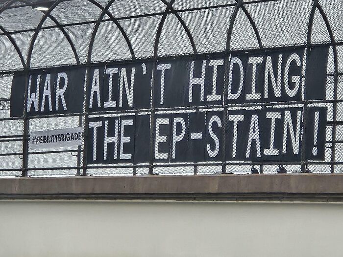 Banner on a fenced overpass with text protesting war and the Epstein scandal in a dystopian United States setting.