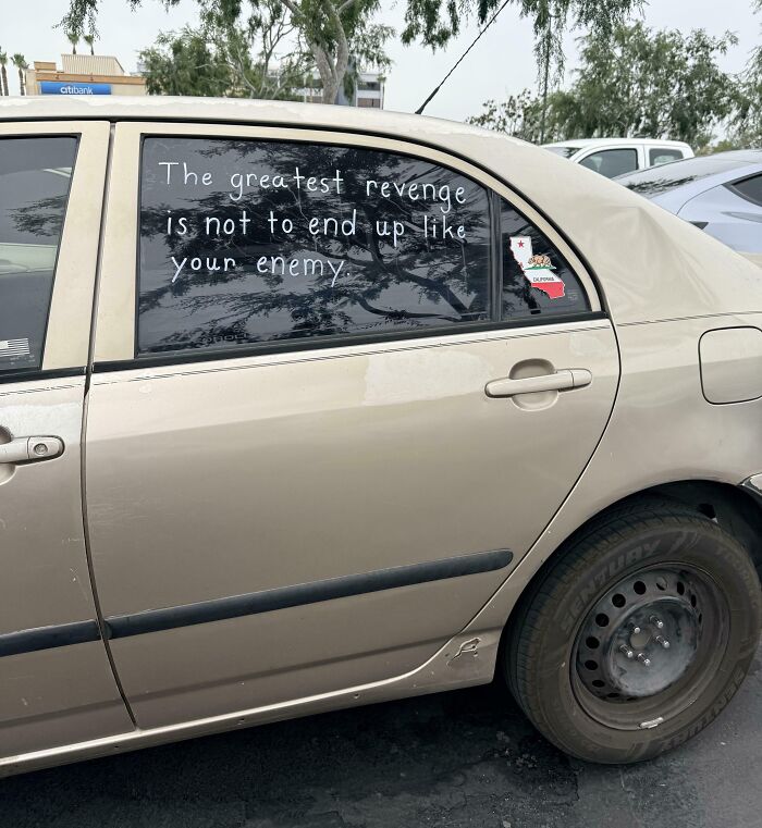 Car window with handwritten quote symbolizing dystopian United States, parked in an urban area with visible wear and tear.