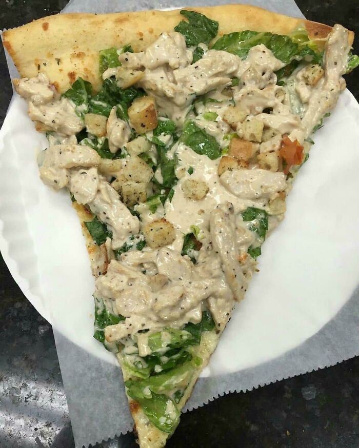 A slice of Caesar salad pizza, a bananasausage food crime, served on a white paper plate.