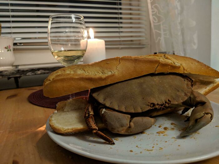 A whole crab in a baguette on a plate with wine and a candle. A Bananasausage food crime.
