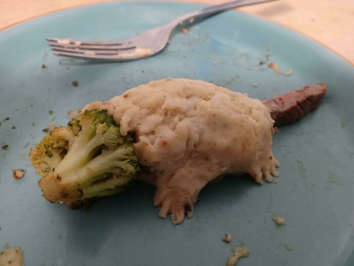 Mashed potato creature with broccoli head and meat tail on a blue plate, a bananasausage food crime.