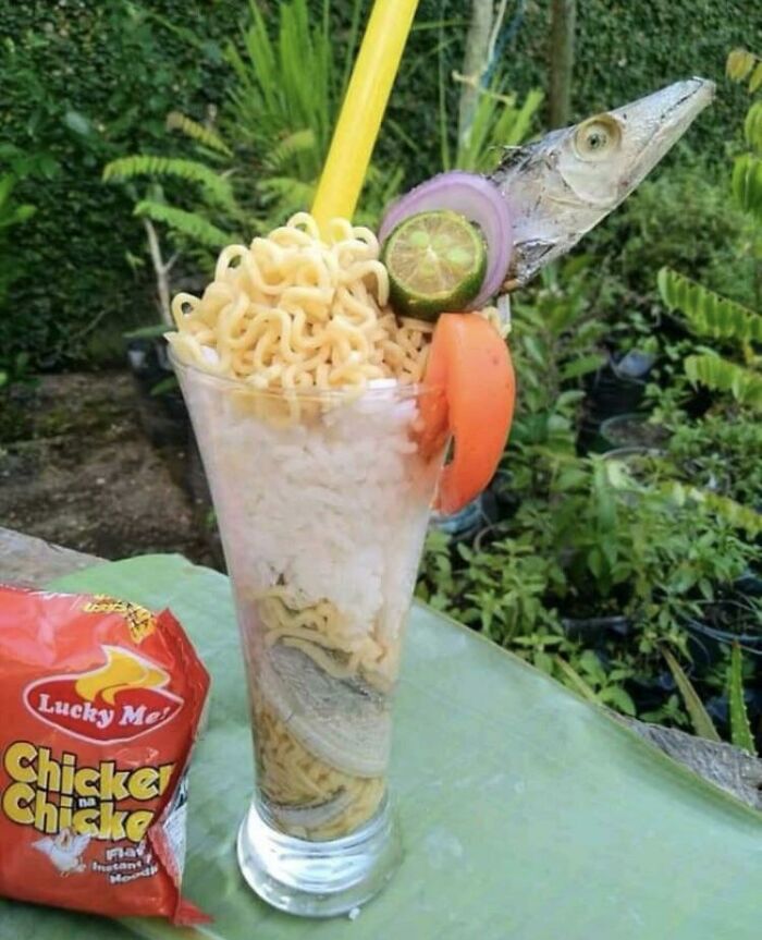 A glass with layered instant noodles, rice, a dried fish head, onion, lime, and tomato. A Bananasausage Food Crime.