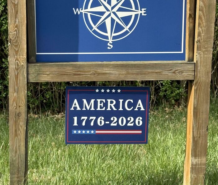Sign with vintage design reading America 1776-2026 in a grassy outdoor setting reflecting dystopian United States imagery.