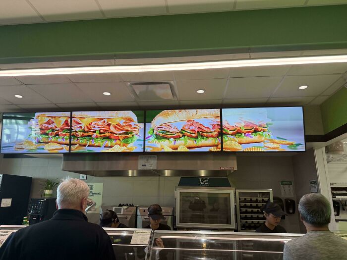Customers standing at a deli counter under digital menus showing sandwiches in a dystopian United States setting.
