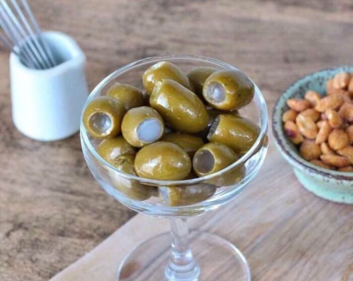 A wine glass full of green olives, some stuffed, next to a bowl of peanuts. A bananasausage food crime.