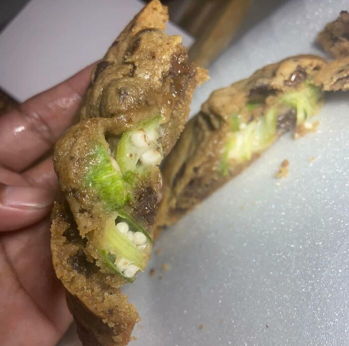 A hand holding a chocolate chip cookie, split open to reveal green okra slices inside. A bananasausage food crime.