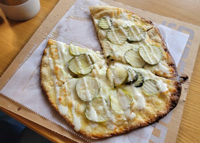 A slice of pickle pizza with white sauce, resembling a Bananasausage food crime, sitting on parchment paper in a cardboard box.