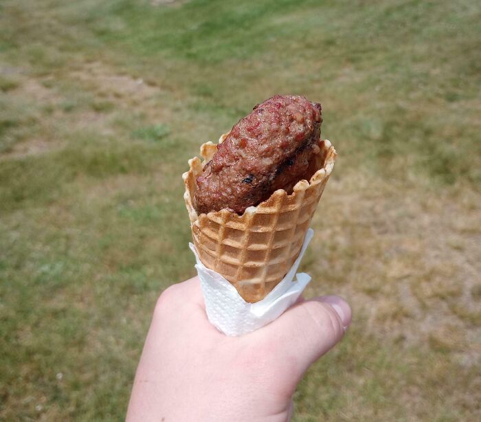 Hand holding a waffle cone with a bananasausage inside, a true food crime, against a grassy background.