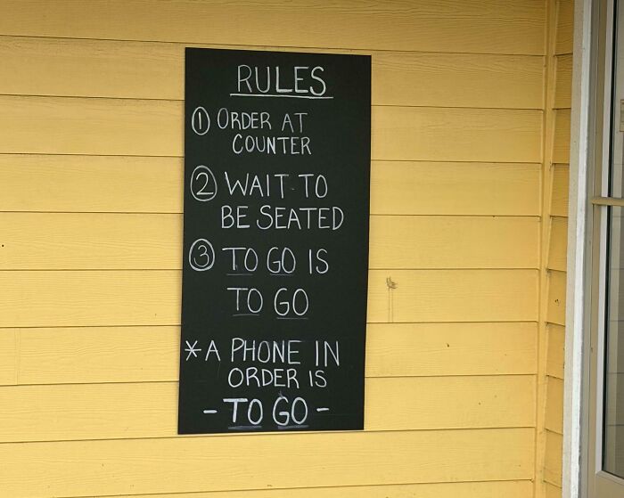 A chalkboard sign with specific rules on a yellow wall, highlighting a rule about to-go orders. It makes you wonder which idiot inspired them.