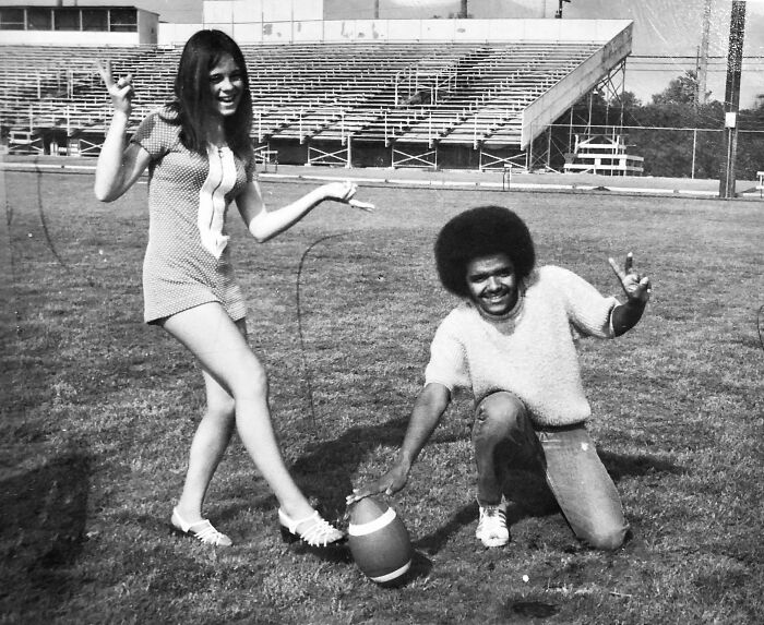 Candid old photo of a young man and woman on a grassy field with a football, both making peace signs.