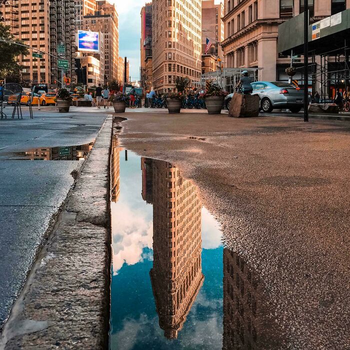 A satisfying picture of the Flatiron Building reflecting in a large puddle on a city street, a gentle reset for your day.