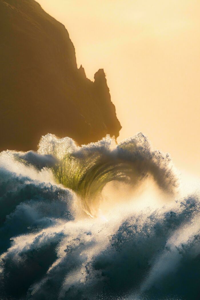 A magnificent ocean wave, backlit by a golden sky, breaks near a dark cliff. A truly satisfying picture.