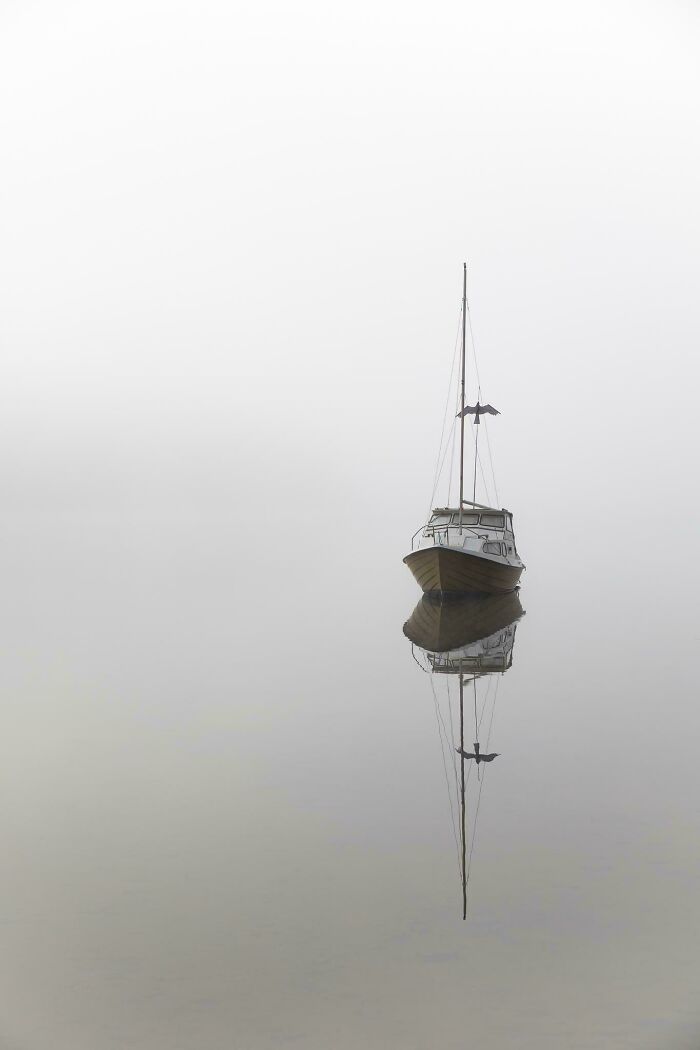 A sailboat calmly floats on a still, foggy lake with its reflection visible, providing a satisfying picture for a reset.