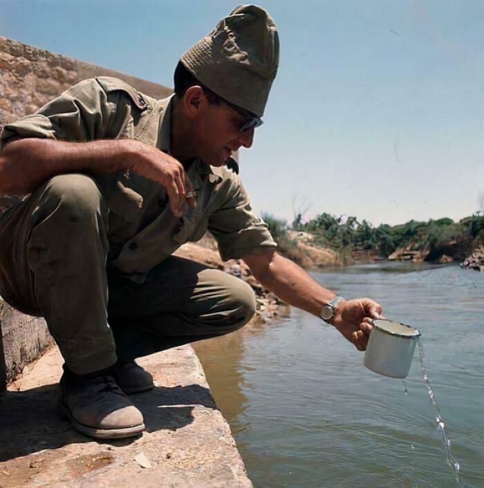 Soldier collecting water in a cup by a riverbank, a historical photo representing a vastly different world from today.