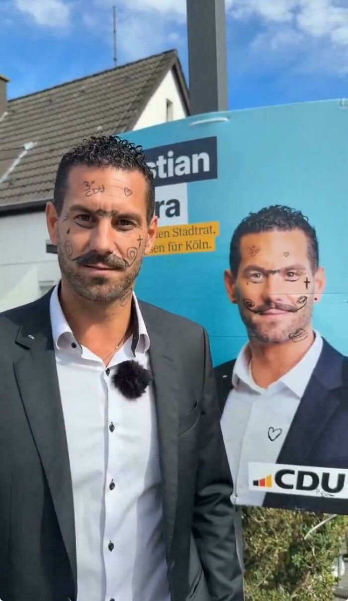 Man in suit with drawn-on face doodles stands in front of a vandalized political poster weird confusing cursed image