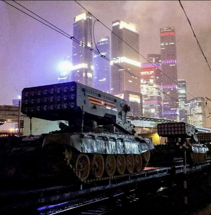 Military tanks on train tracks with city skyscrapers glowing in the background