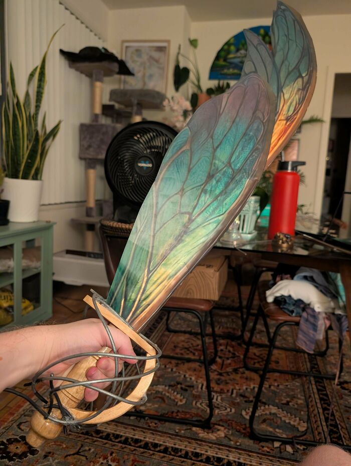 A hand holds a handmade fantasy sword with iridescent, butterfly-like wings for the blade, a unique craft.