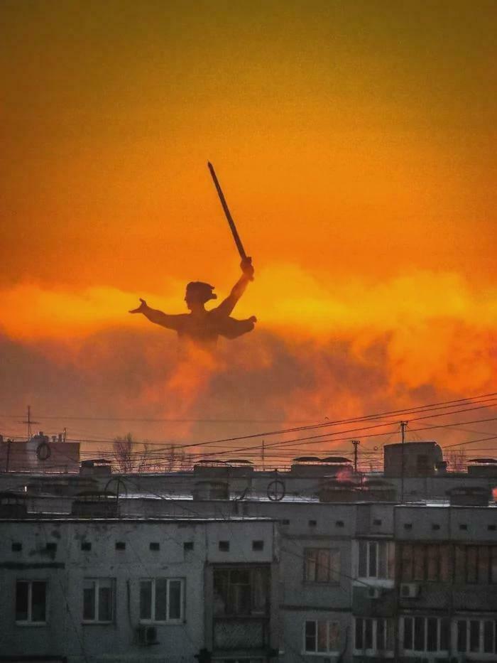 Silhouette of a statue with sword over urban buildings during eerie orange sunset