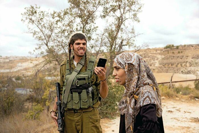 A man in military fatigues with an unfortunate haircut shows a phone to a woman in a hijab.