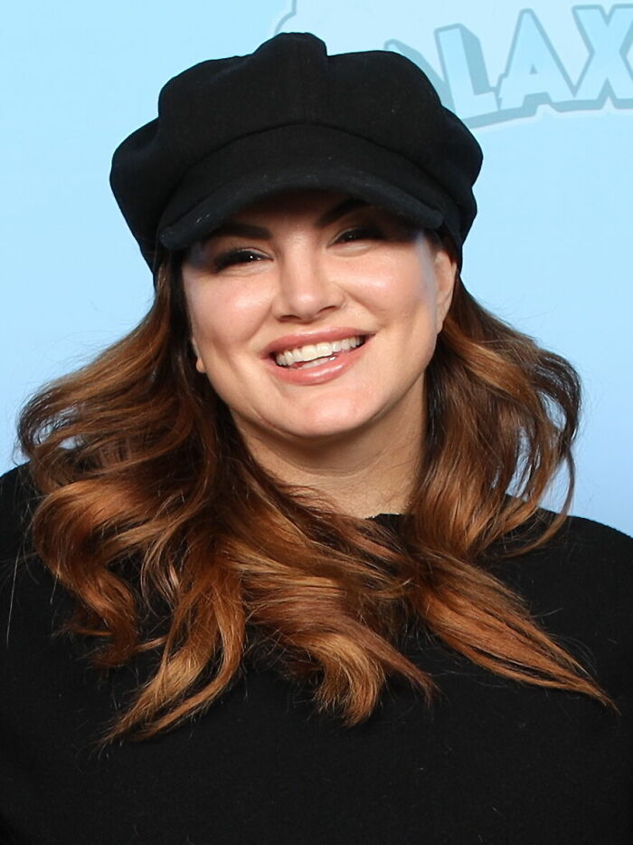 Gina Carano smiling, wearing a black hat and black top, showcasing her career highlights in a light blue background event.