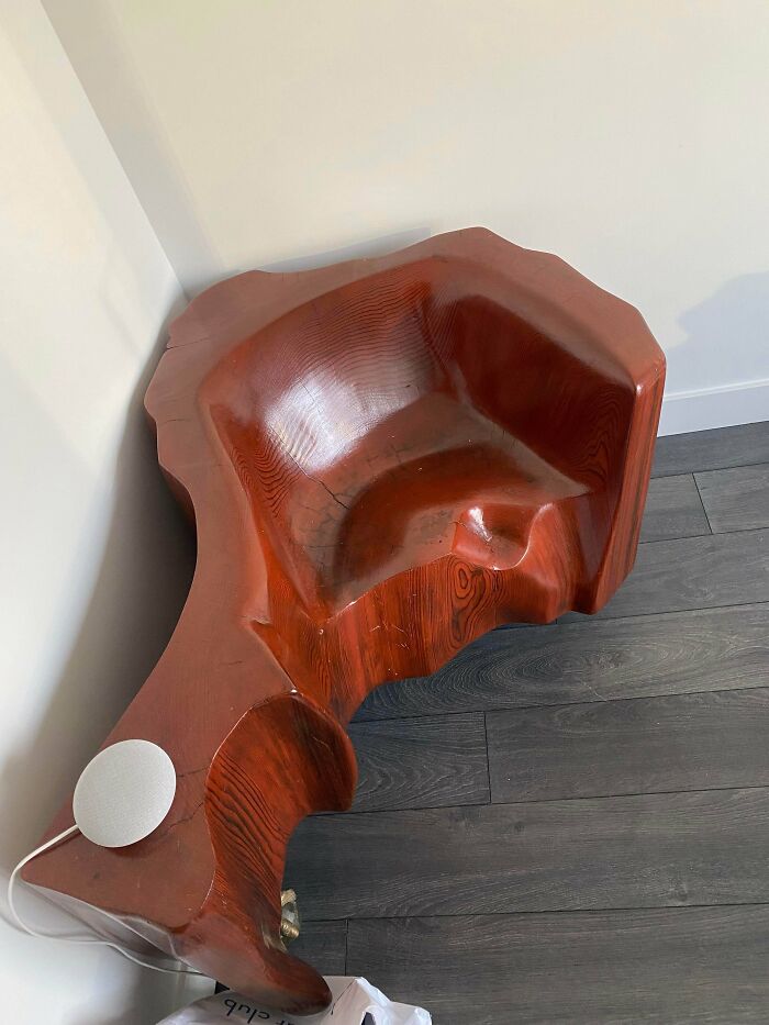 A large, unusual possession: a sculpted, reddish-brown wooden chair with a curvy design, next to a white wall and dark wood floor.